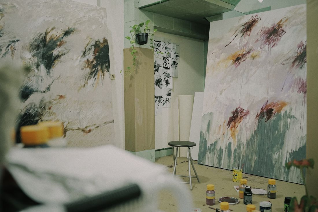 A working studio corner with large abstract paintings on the walls, smaller ink studies pinned beside them, and paint jars on the floor