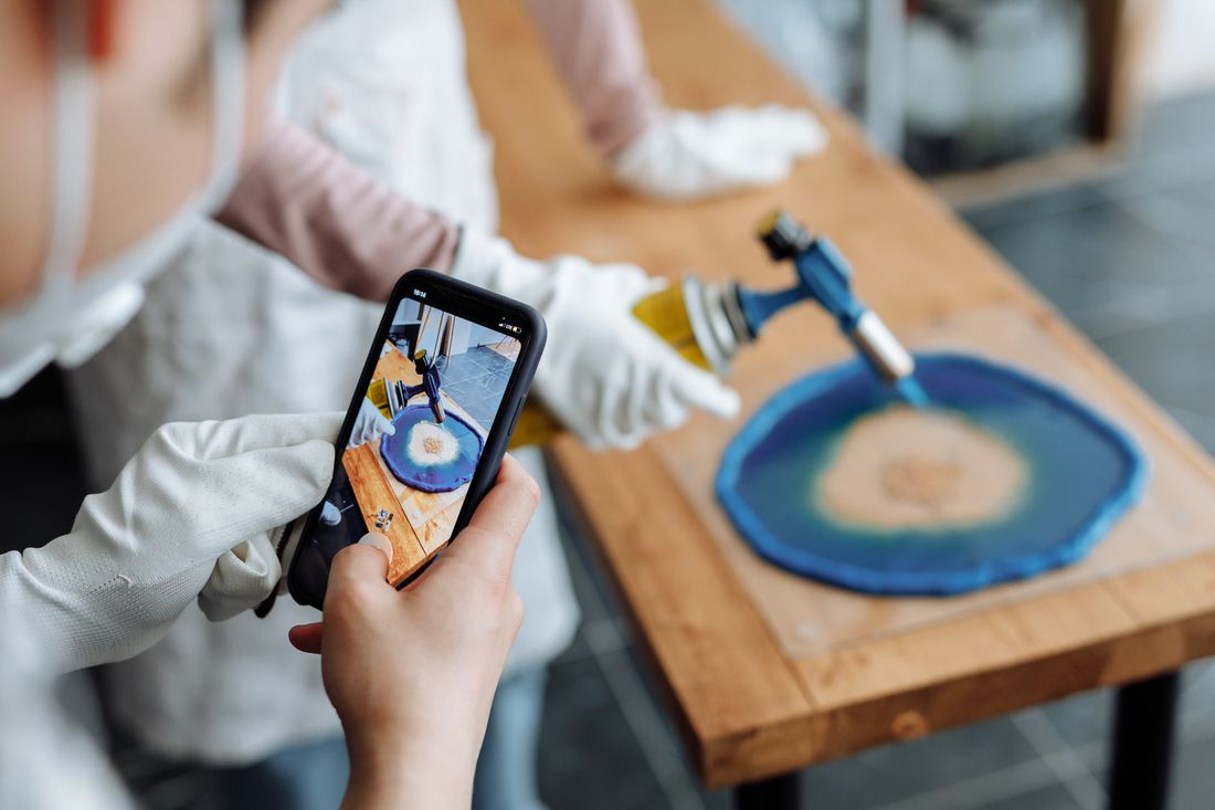 A gloved hand photographing a resin art piece on a smartphone while it's finished with a small torch