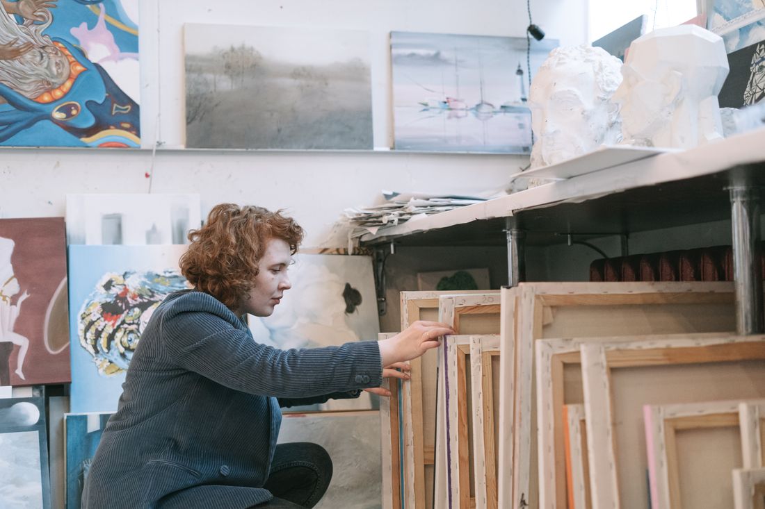 An artist crouches beside a stack of unframed canvases leaning under a studio shelf, sorting through them by hand while more finished paintings line the wall above.