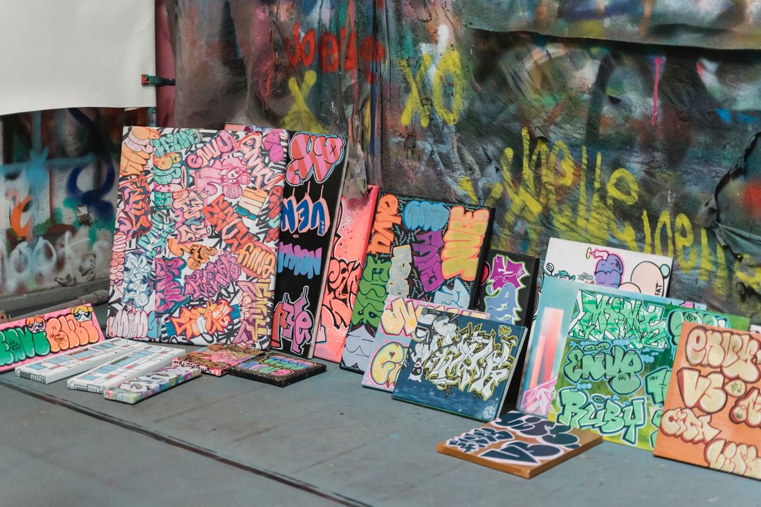 A row of brightly painted graffiti-style canvases of different sizes leaning against a paint-covered studio wall, with smaller panels resting on the floor in front of them.