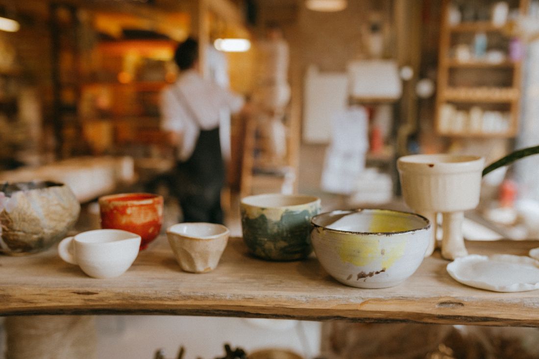 A row of handmade ceramic cups and bowls in different glazes lined up along a wooden workshop shelf, with a person working in the blurred background.