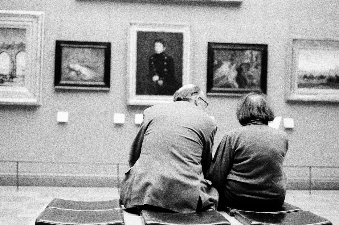 An elderly couple sitting together quietly looking at framed paintings on a gallery wall