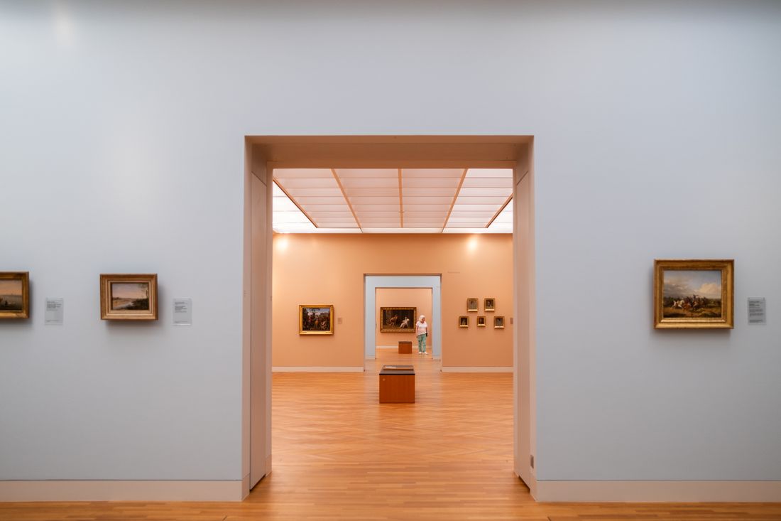 A quiet, empty museum gallery seen through an open doorway, with framed paintings on the walls and one distant visitor