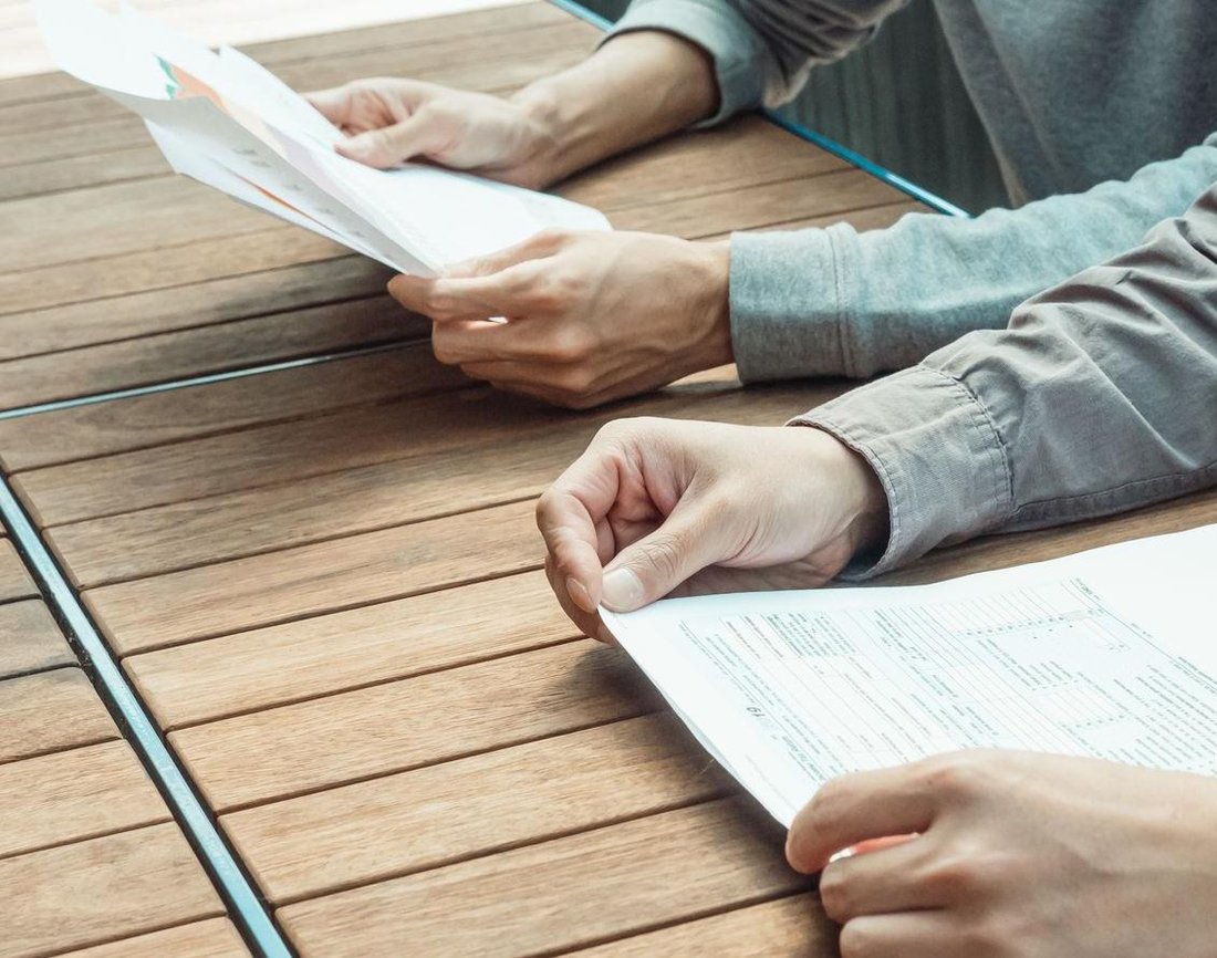 Two people at a table each hold a printed sheet of paper, comparing pages side by side in daylight.