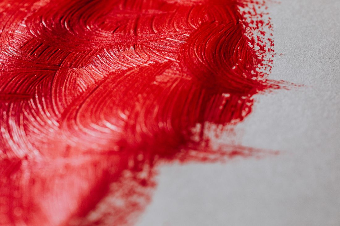 Close-up of thick red paint brushstrokes on a light painted surface, raking light bringing out the ridges and texture.
