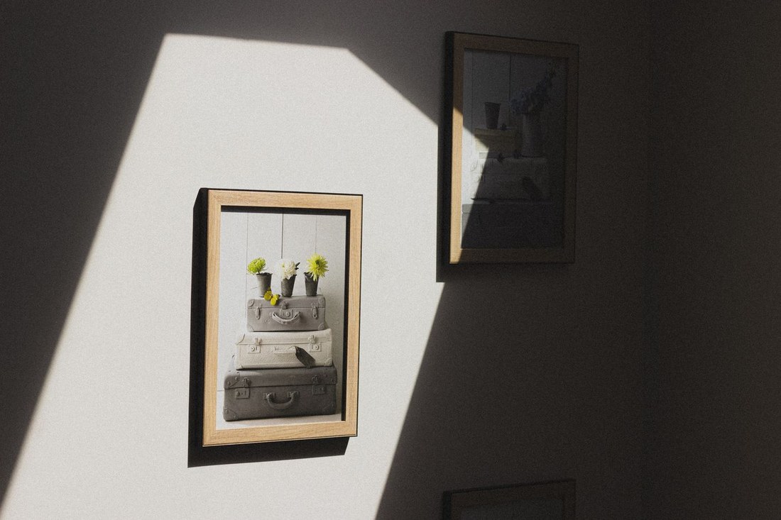 Two framed pictures hung on a plain pale wall, with a diagonal band of window light crossing it and the rest of the wall in shadow.