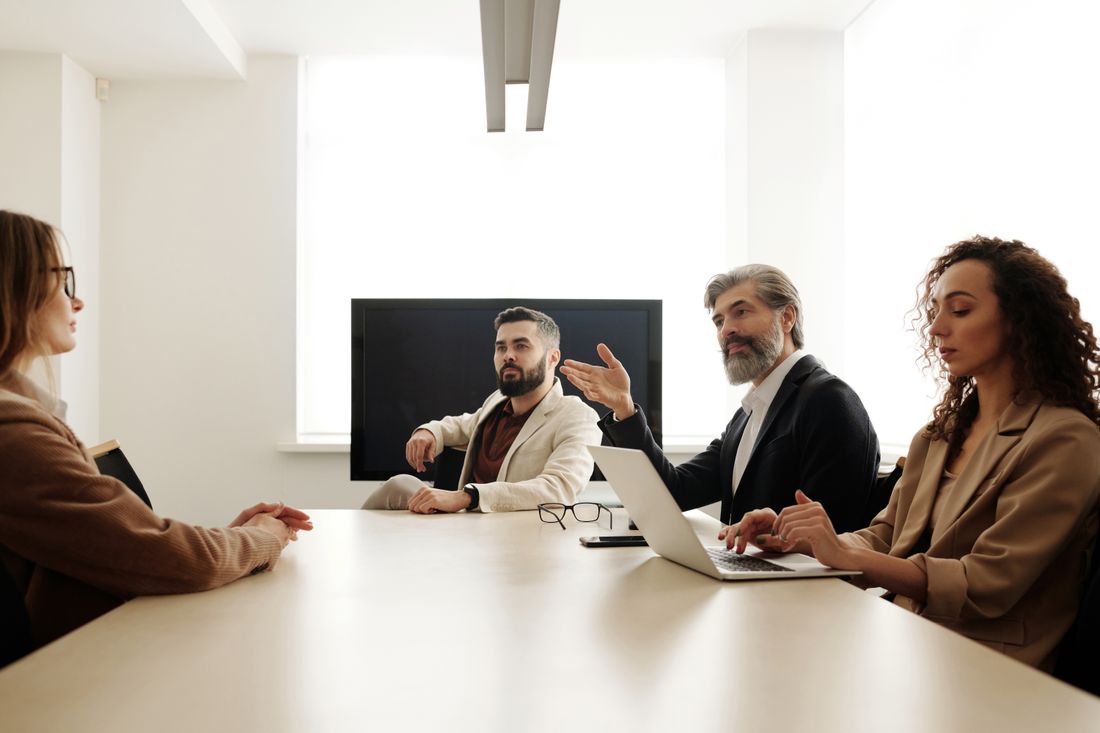 Four colleagues seated around a plain white table in a bright meeting room, one man gesturing while speaking, a blank screen behind them.