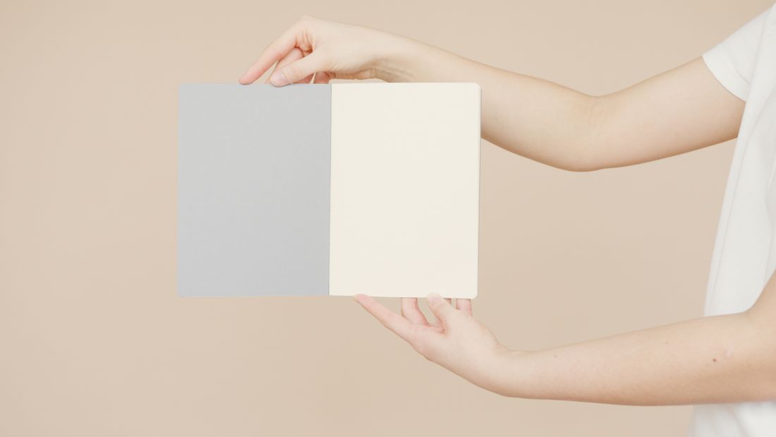 Two hands holding open a blank notebook at an angle against a plain beige background, no text visible.