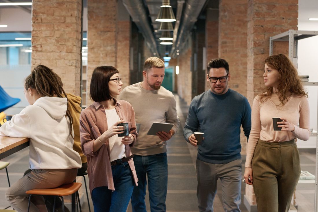 A small group of coworkers talking together in a plain brick-walled corridor, holding coffee cups.