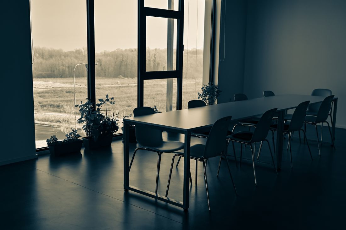 An empty meeting room with a long table, stacking chairs and daylight from a full-height window.