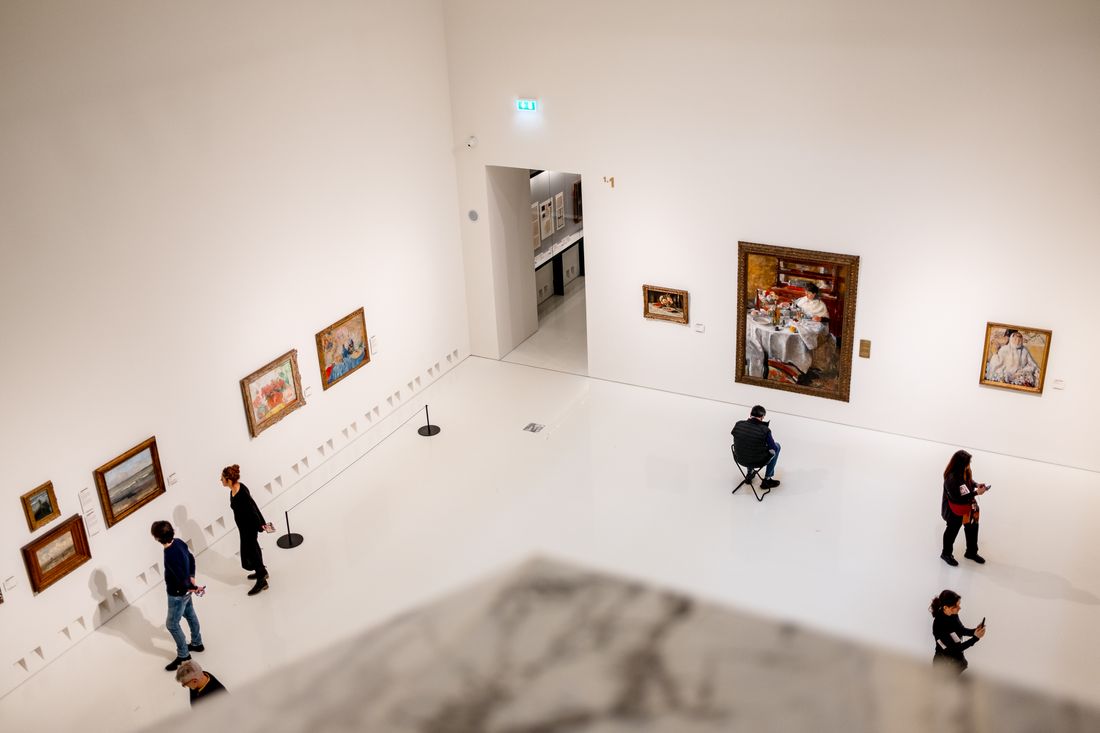 A high-angle view of visitors walking through a white-walled gallery room hung with framed paintings