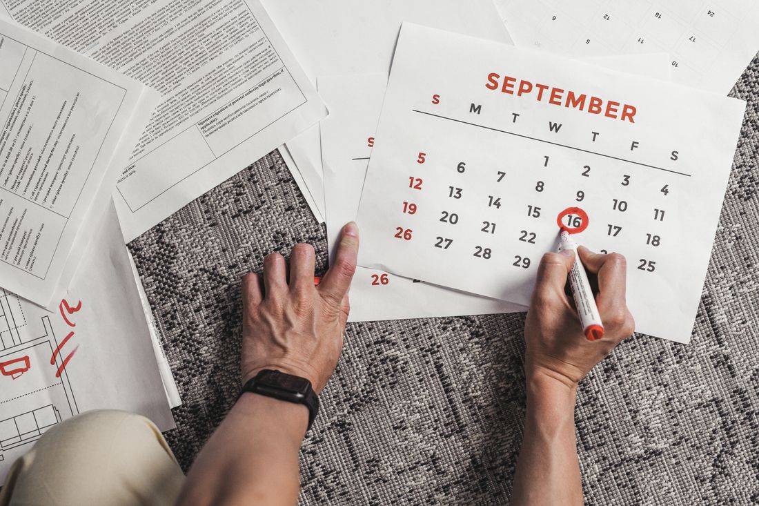 A hand circling a date on a paper calendar with a marker, papers scattered nearby