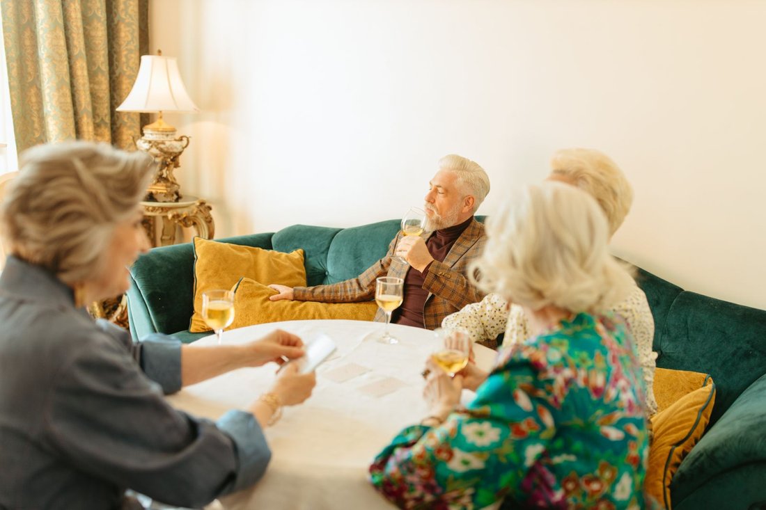 Four older adults sit close together around a low table on a green velvet sofa in a warmly lit room, glasses of wine and a few face-down cards on the table between them.