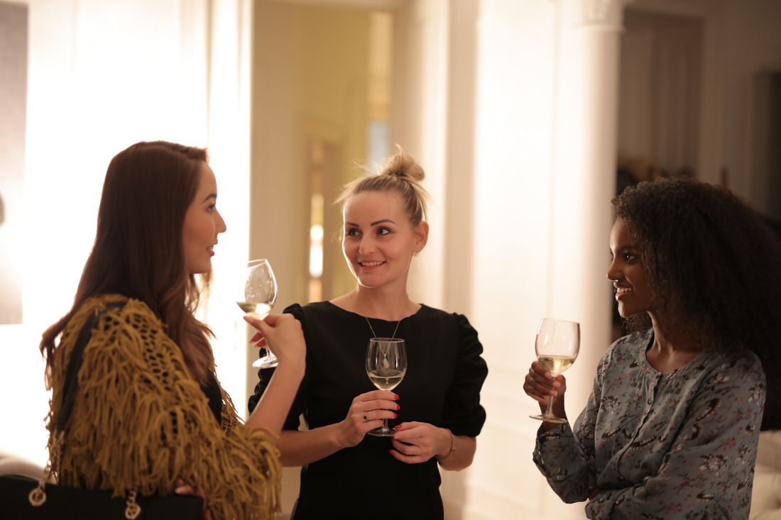 Three women stand together in a warm, softly lit room with a white column behind them, each holding a wine glass and talking mid-conversation, seen from the side.