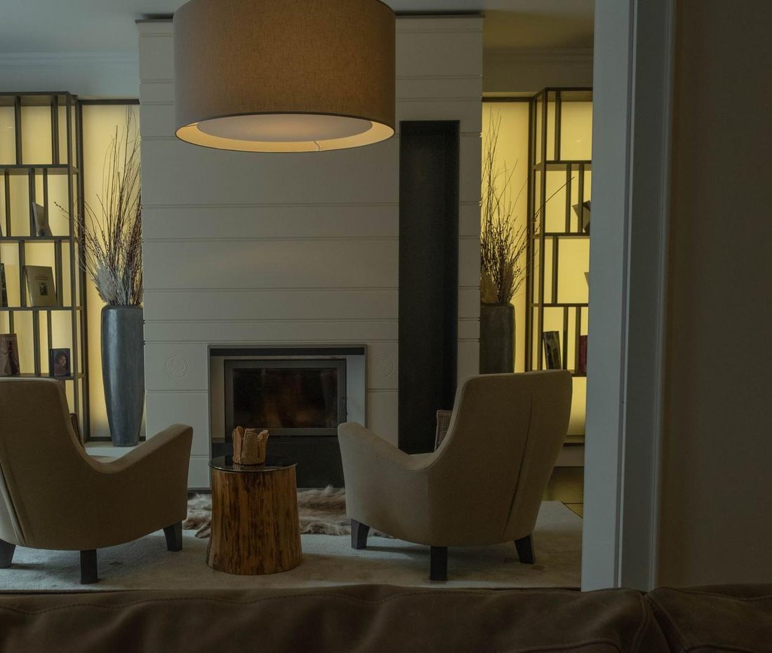 Two empty armchairs and a low wooden stump table arranged in front of an unlit fireplace in a warmly lit room, with a lamp glowing above and nobody in the room.