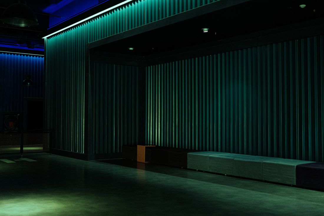 An empty event room lit in green and blue, with slatted walls, low benches along one side and a bare polished floor before an audience arrives.