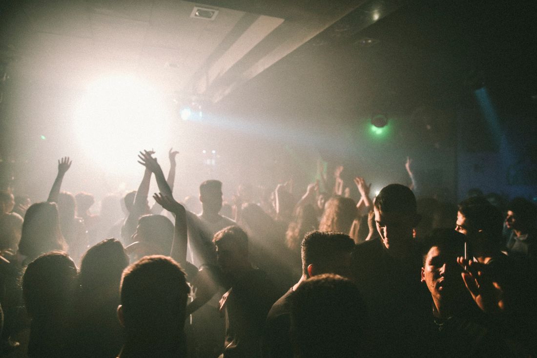 A hazy indoor crowd with arms raised, silhouetted against a bright white backlight with a touch of green colored light in the background.