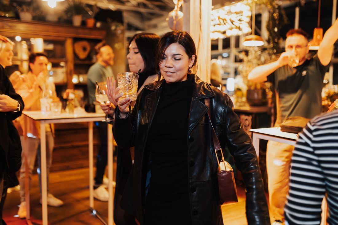 A woman holding a cocktail glass amid a warmly lit indoor social gathering, other guests talking and drinking nearby.