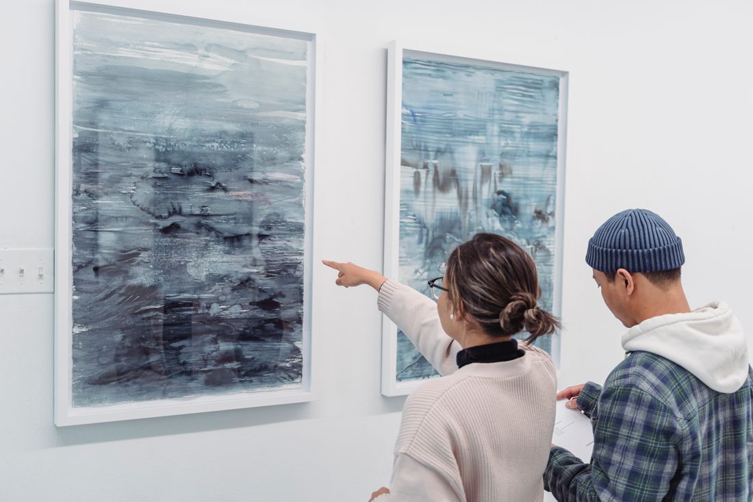Two people stand close to a pair of framed abstract works on a white gallery wall, one of them pointing at the left picture while the other looks on.