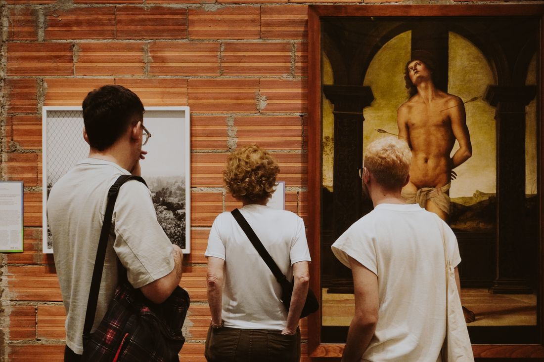 Three visitors stand side by side facing framed pictures hung on an exposed brick wall in a gallery, seen from behind.