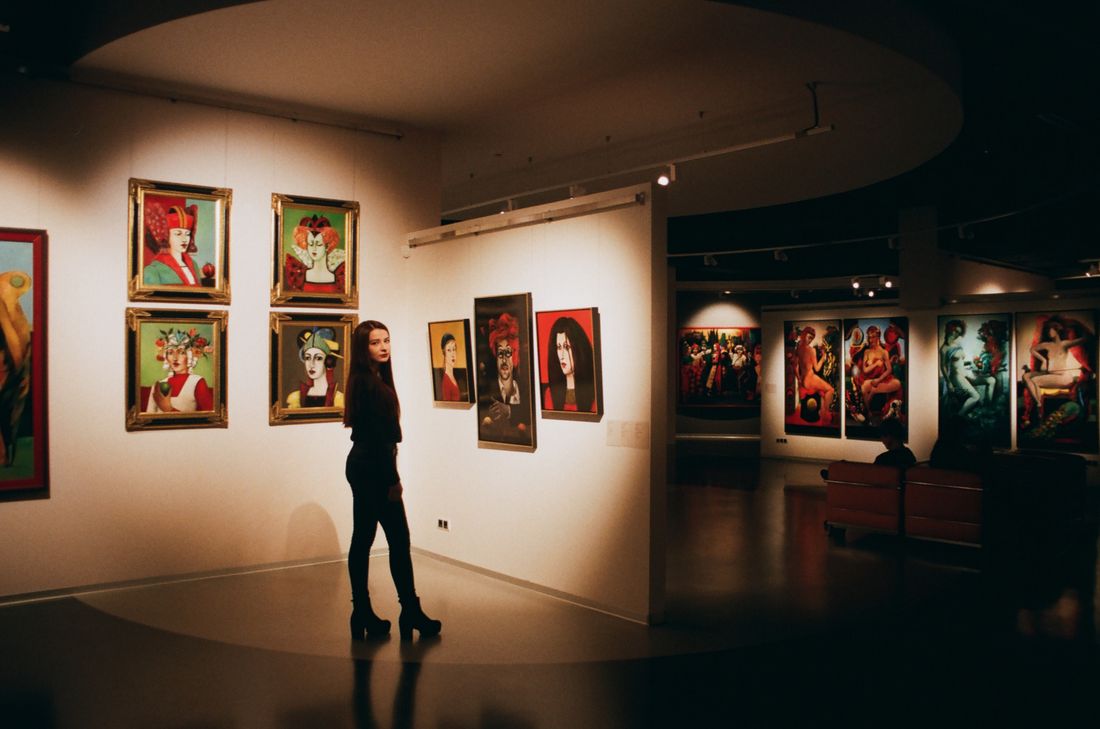A wide view of a dimly lit gallery room with framed figurative paintings along a curved white wall and a single visitor standing in front of them.