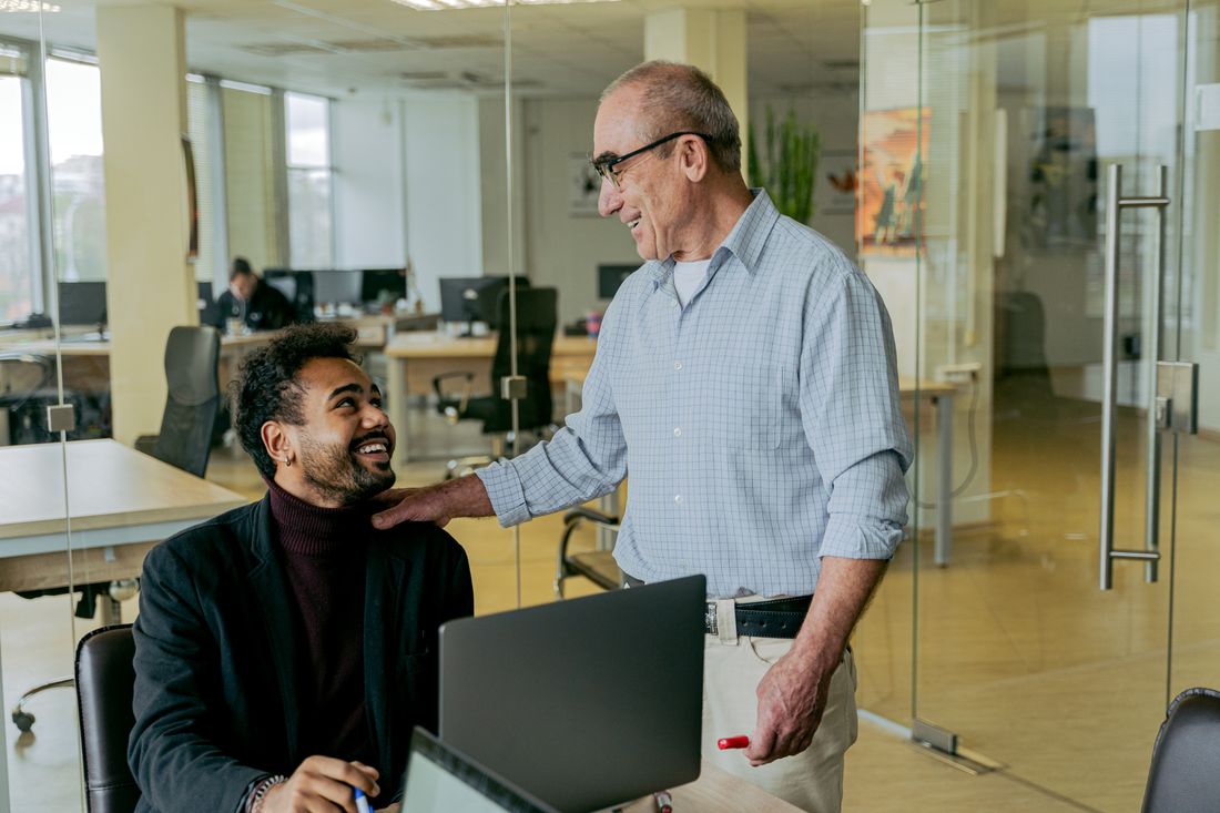 A staff member greets an arriving visitor at a desk with a laptop, both smiling mid-conversation