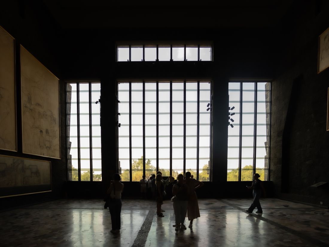 Visitors silhouetted against a tall bank of windows in a dim museum gallery, daylight streaming in