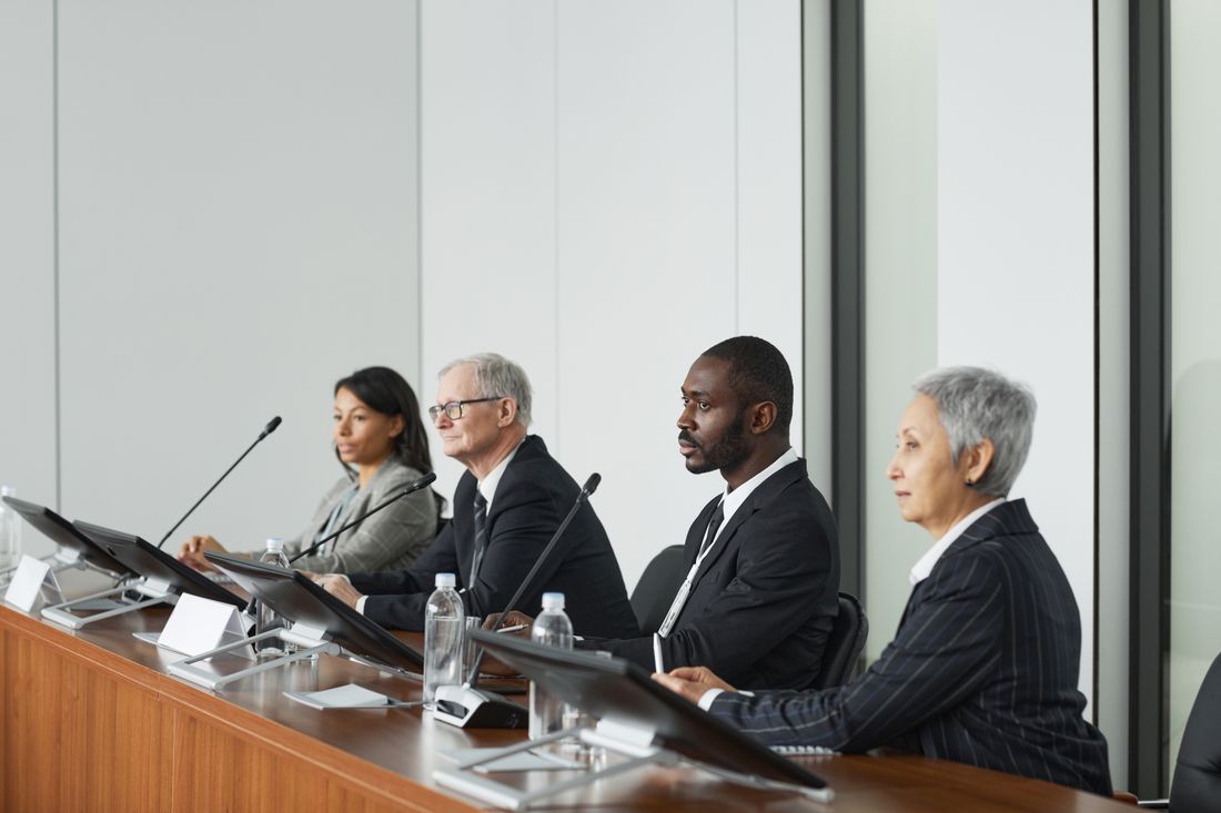 Four panelists seated in a row at a conference table, each with a microphone and name placard