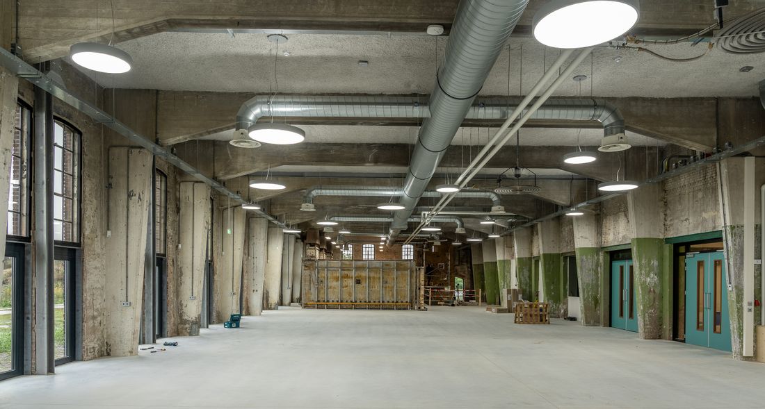 An empty concrete warehouse interior with exposed metal ductwork and large windows, the kind of raw space sometimes used for a temporary exhibition.