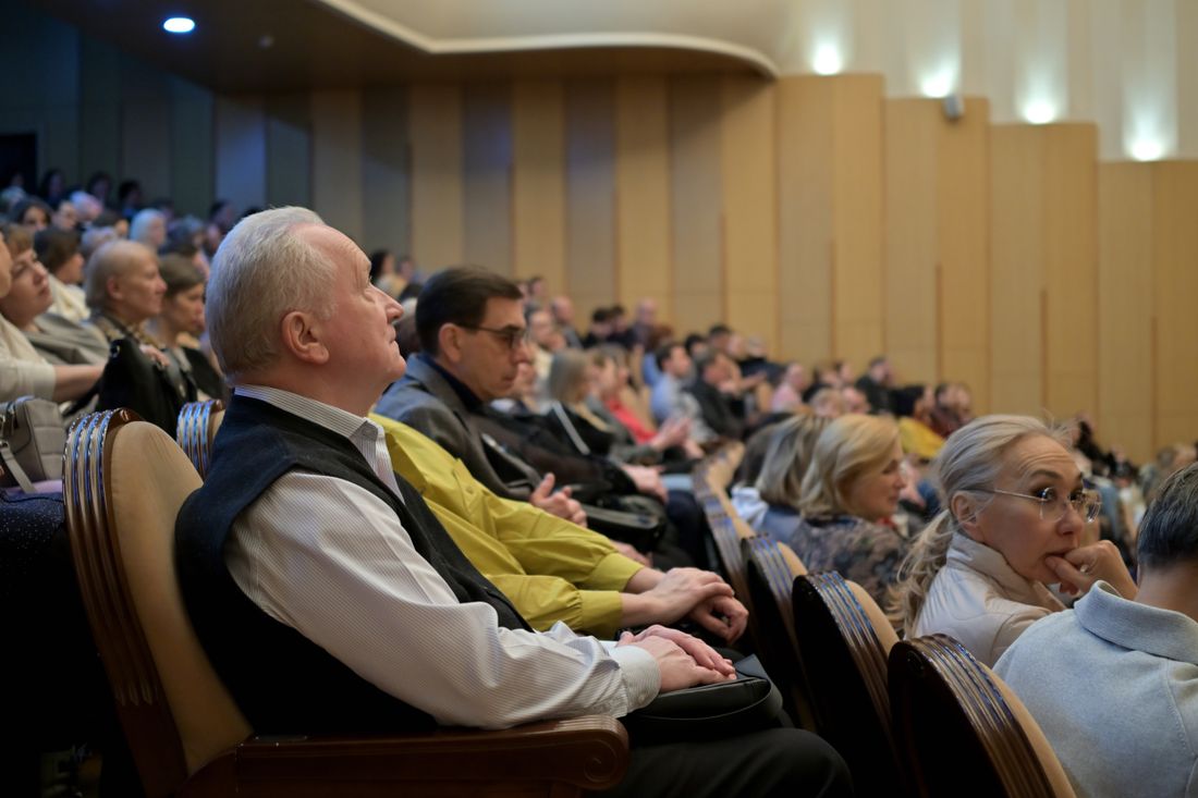 An audience seated in rows in a wood-paneled hall, listening toward the front of the room