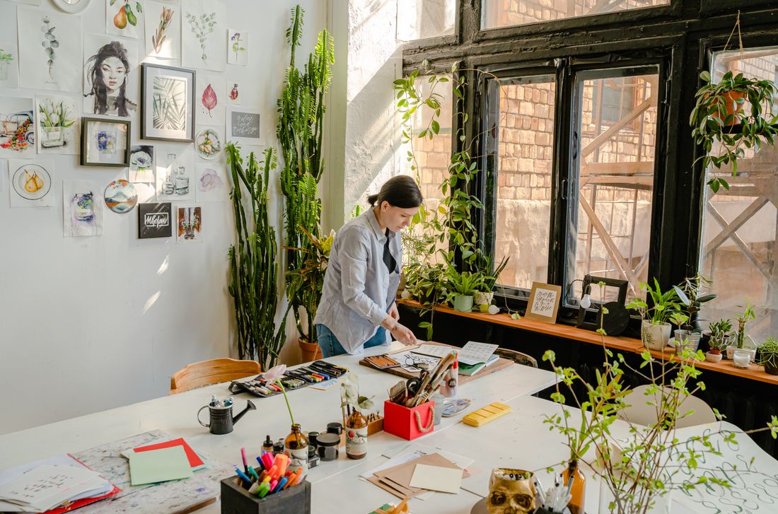 A bright loft studio with drawings and small works covering one wall and a person working at a long table by tall windows