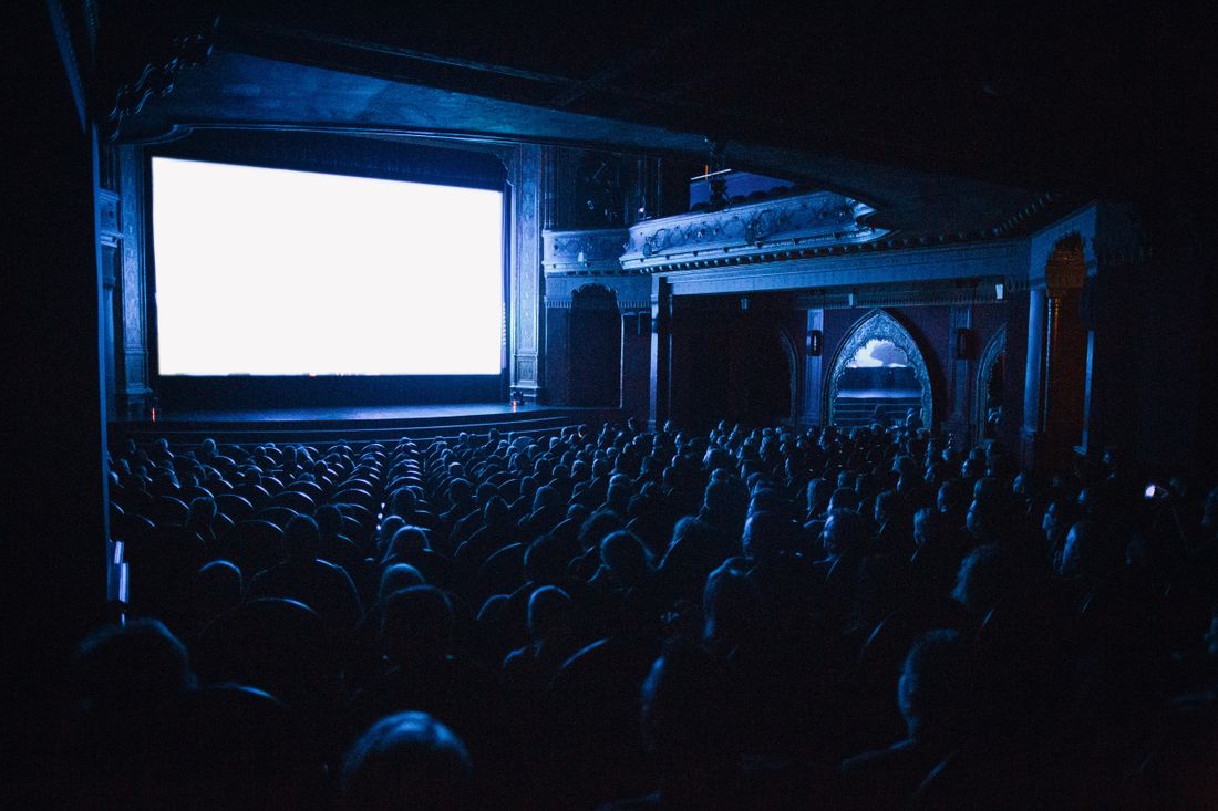 A large darkened theater audience seen from behind, silhouetted heads facing a brightly lit blank screen at the front of the room.