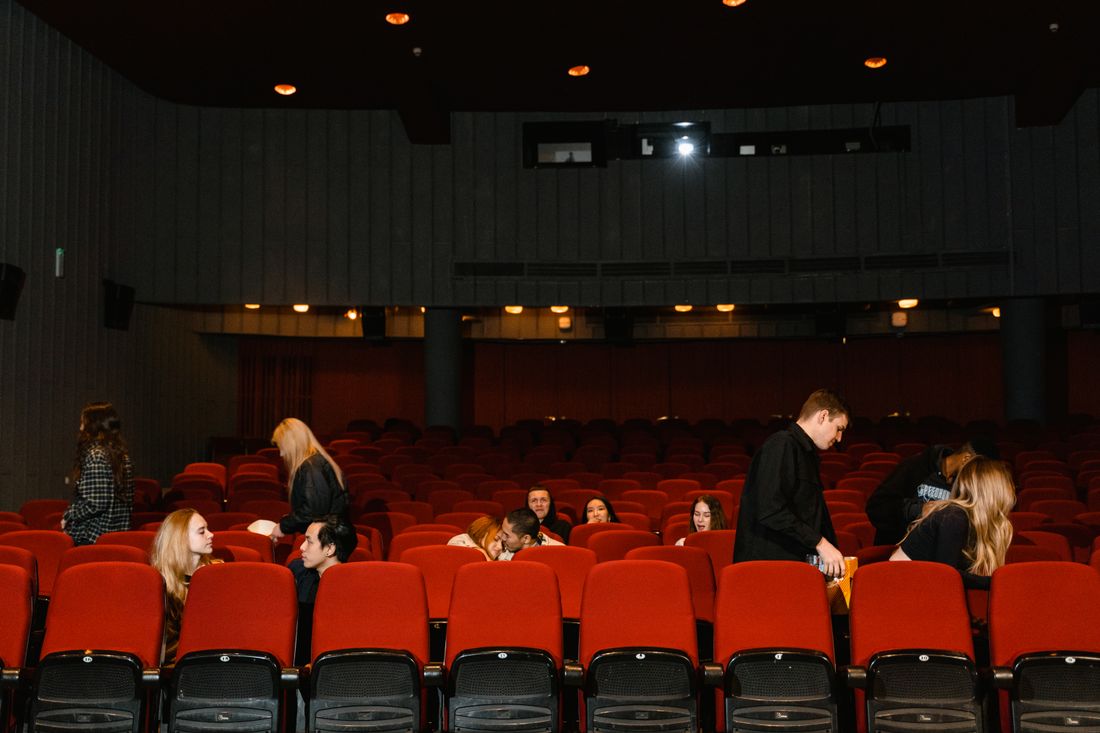 People finding their seats among rows of red theater chairs while the overhead lights are still on before a screening begins.