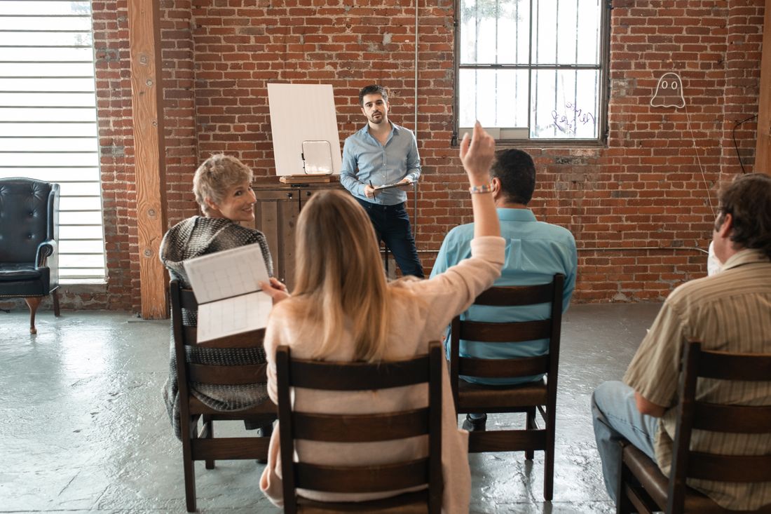 A small seated audience in a brick-walled room, one person raising a hand to ask a question while a presenter stands near a blank easel.