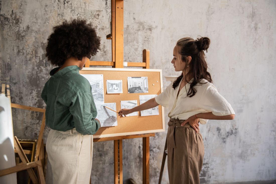Two people seen in profile in a plain studio, one gesturing while explaining pinned images to the other.