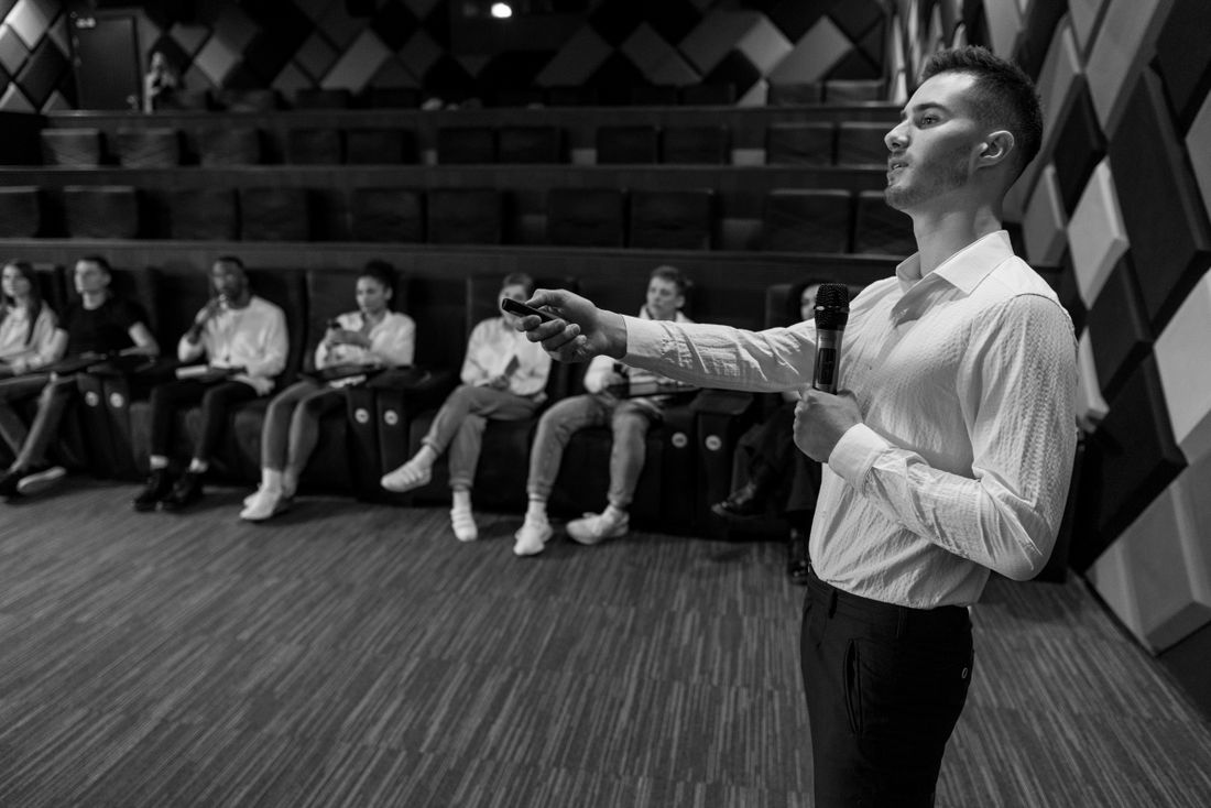 Speaker addressing a seated audience in a patterned indoor lecture room.