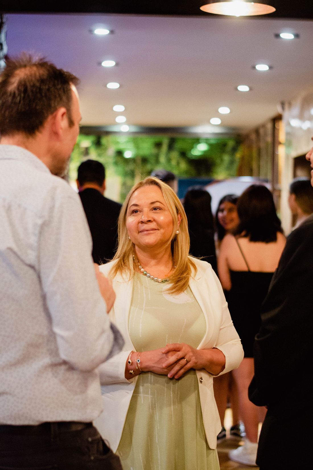 Adults greeting and talking with one another inside a busy indoor gathering.