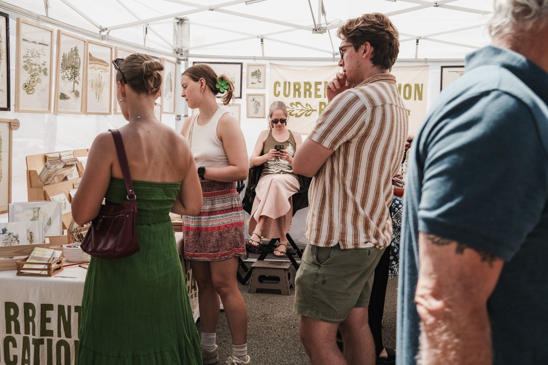 Visitors browsing framed artwork inside an open-sided fair booth under a canopy tent
