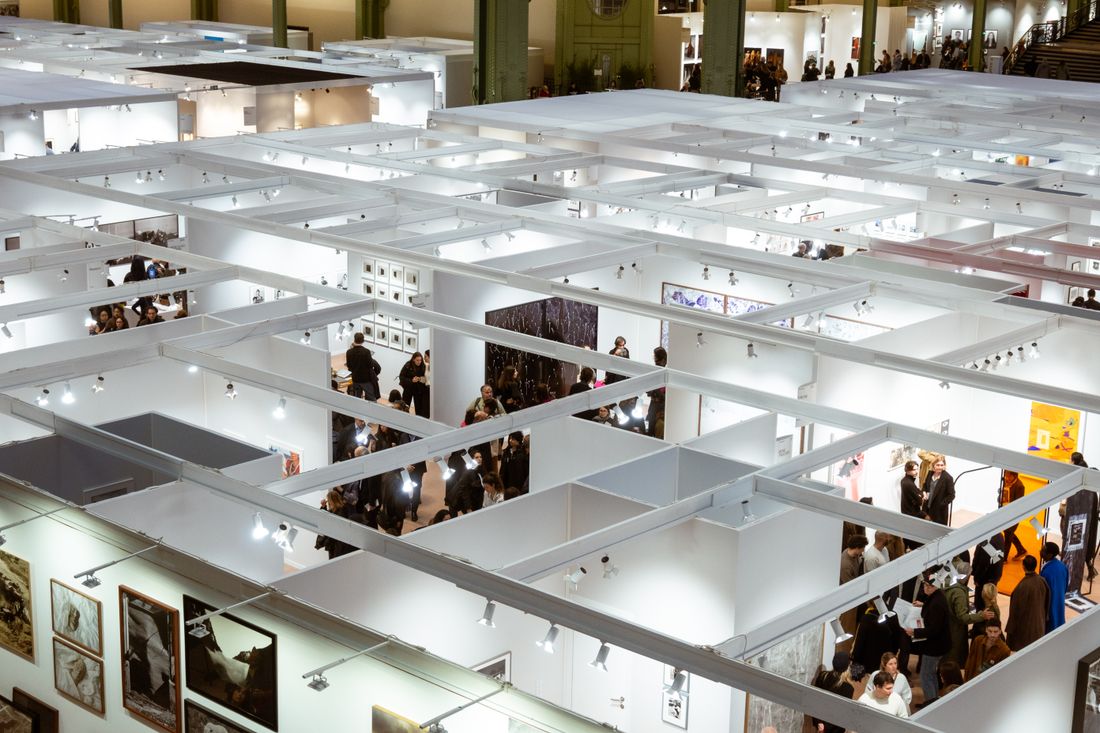 Aerial view of a busy art fair floor with white booth walls, framed artwork, and visitors moving through the aisles