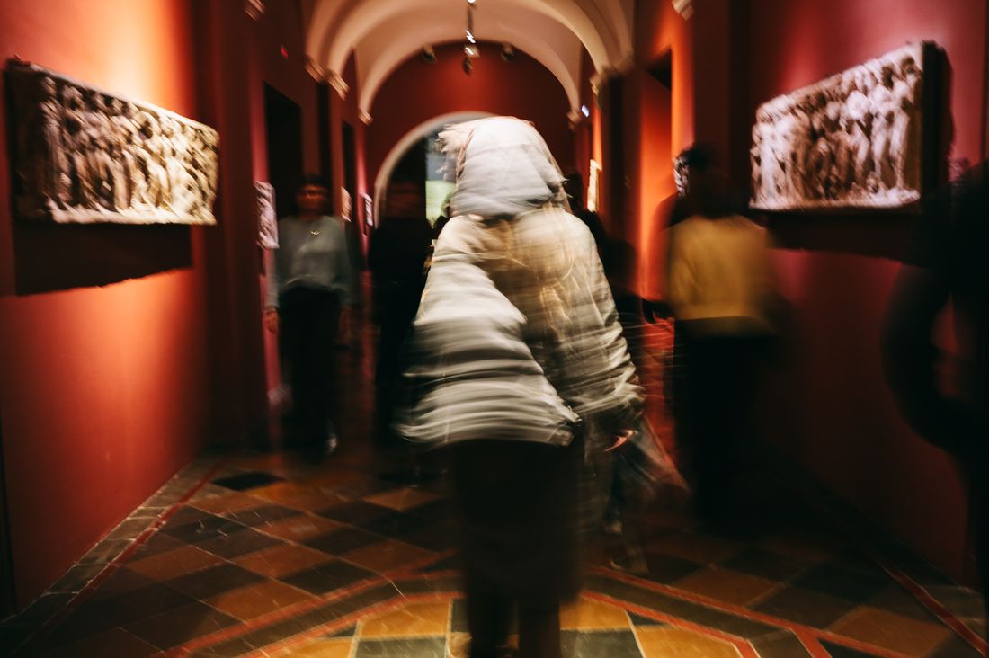 A blurred crowd of visitors moving through a warmly lit, arched gallery corridor with work hung on both walls