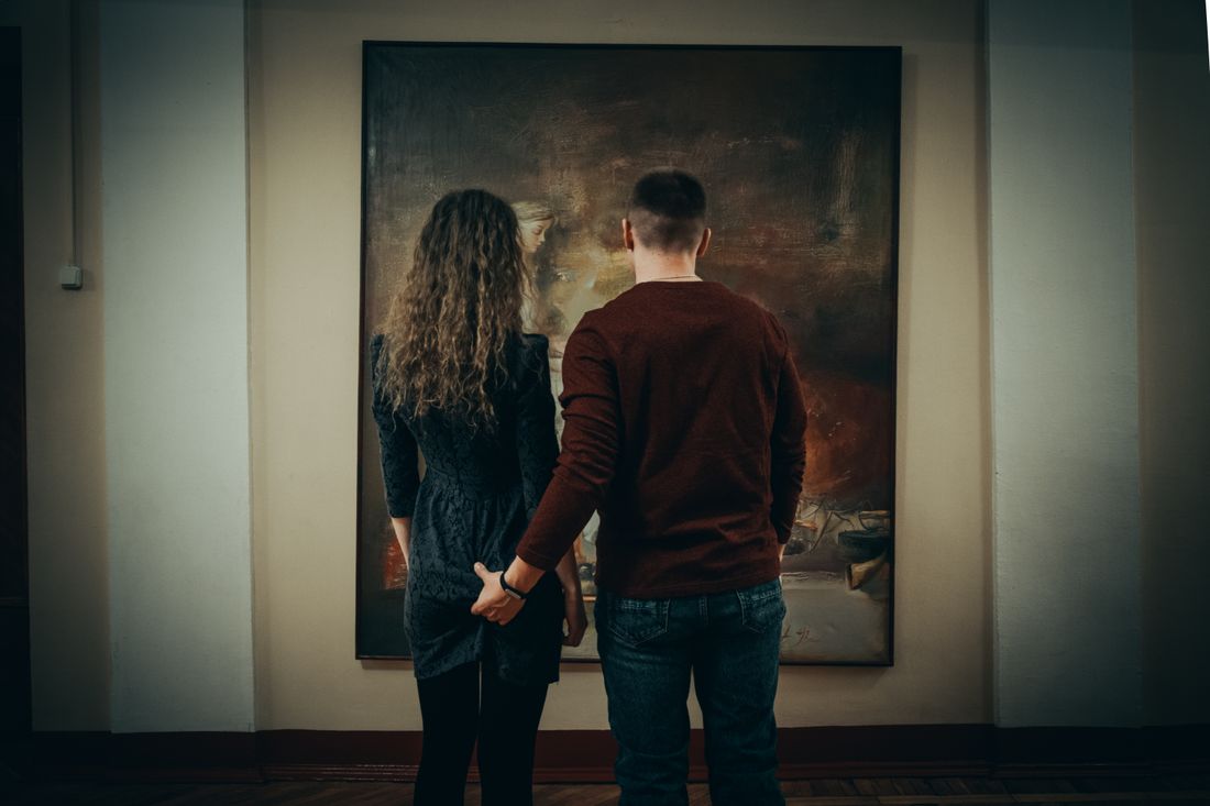 Two people standing close together from behind, hand in hand, looking at a large painting on a gallery wall