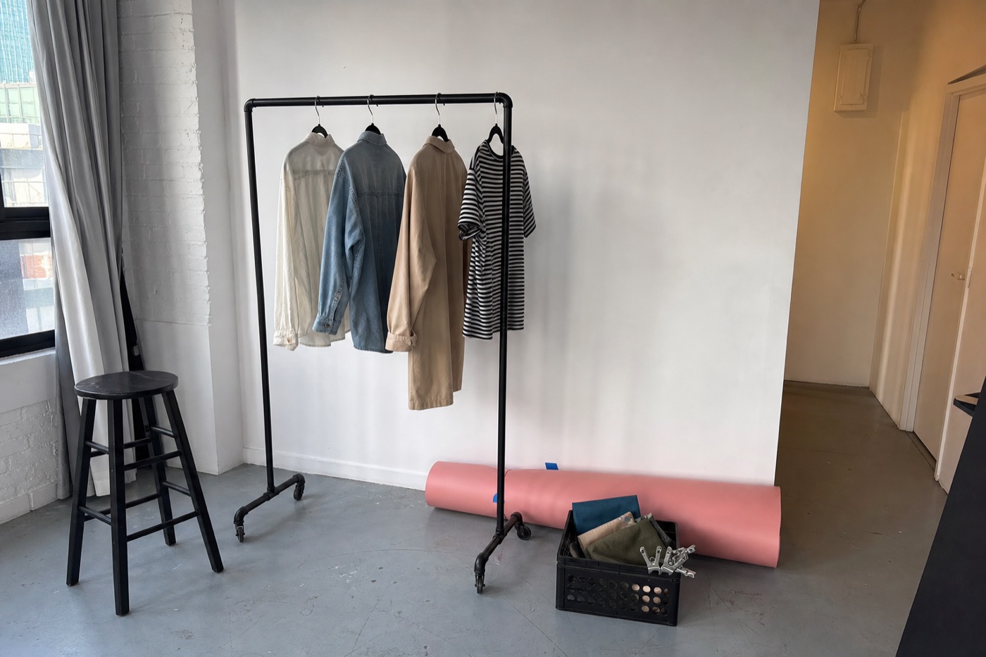 Casual modern studio corner with a supported clothing rack, varied garments, stool, backdrop roll, and crate of fabric samples