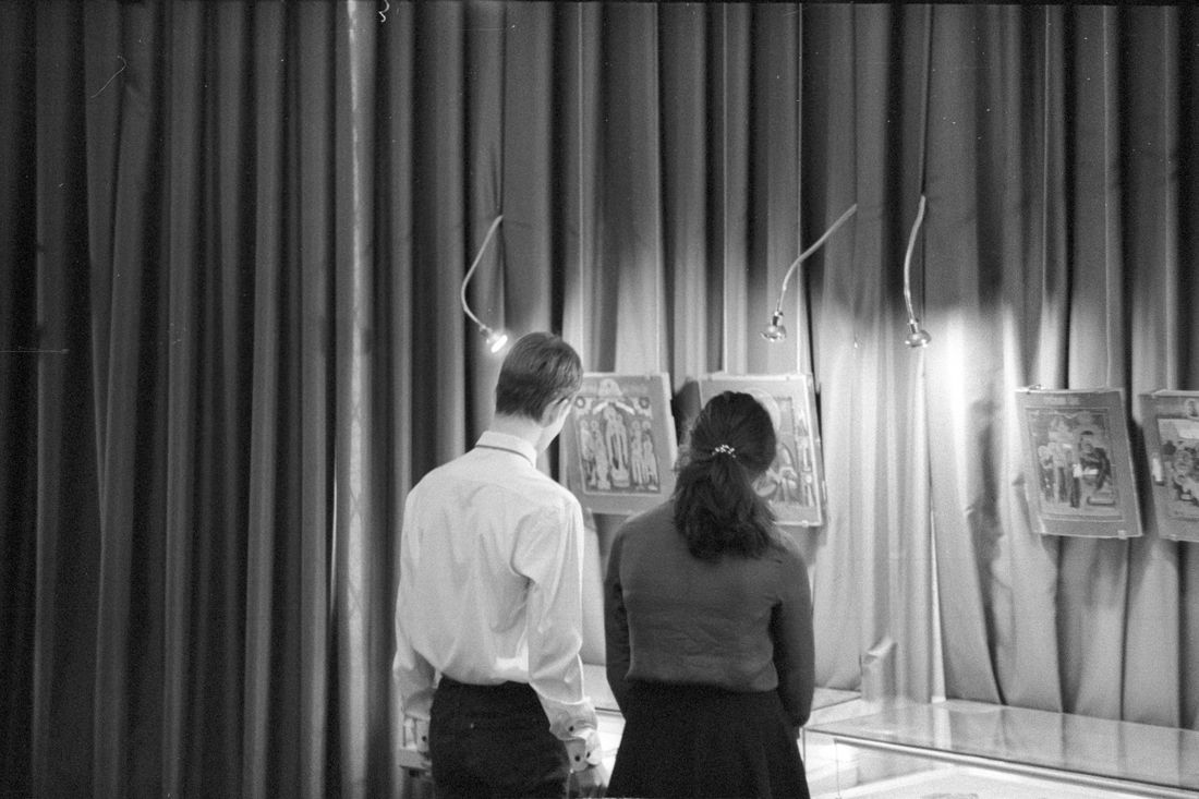Two gallery visitors seen from behind viewing framed artwork in a dim exhibition space