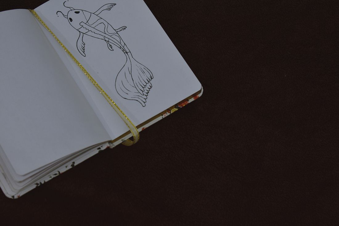 A small sketchbook held open with an elastic band, showing a pencil sketch on the page against a dark background
