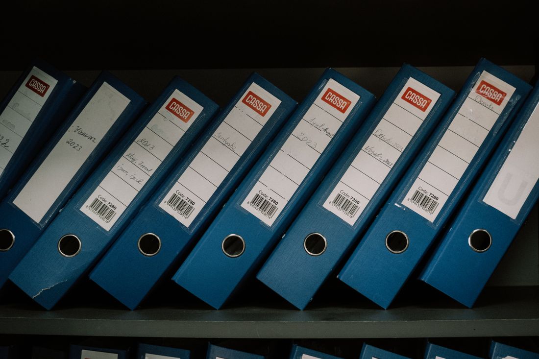 A row of labeled blue binders organized on an office shelf, by date and name