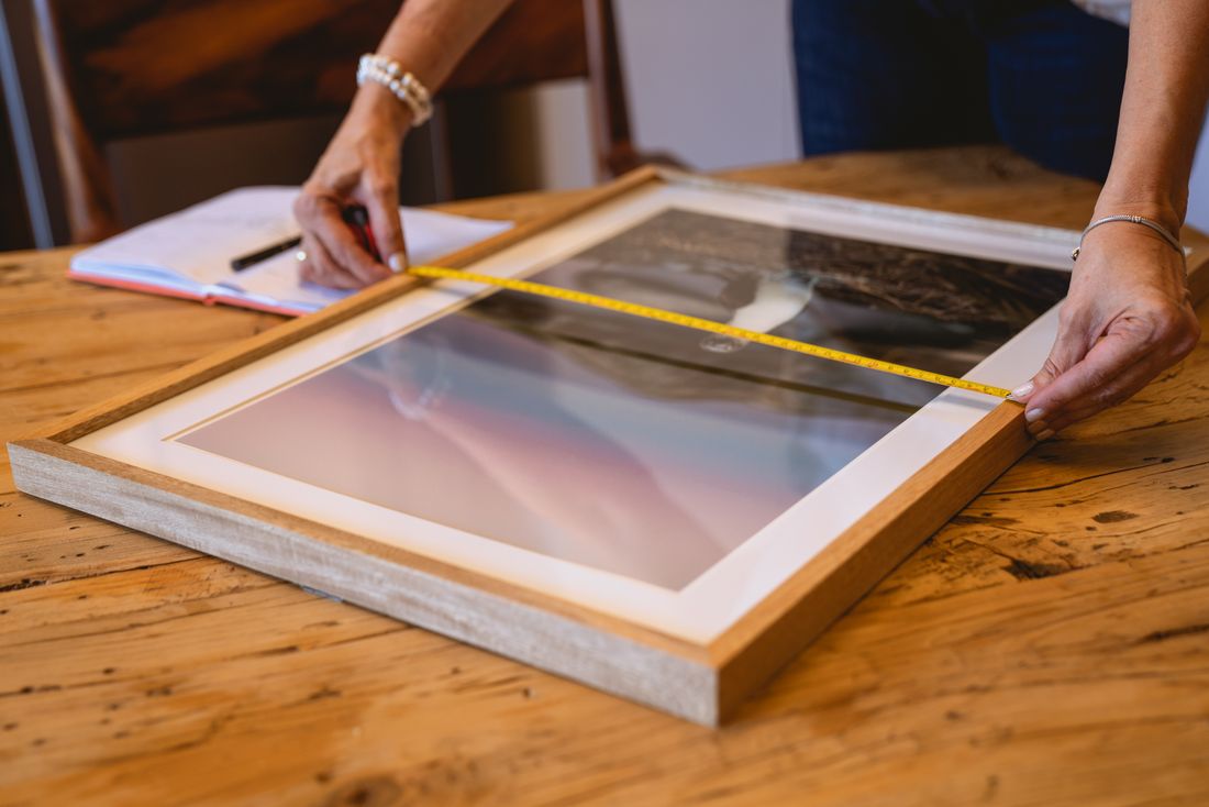 Hands using a tape measure to check the size of a framed print on a wooden table
