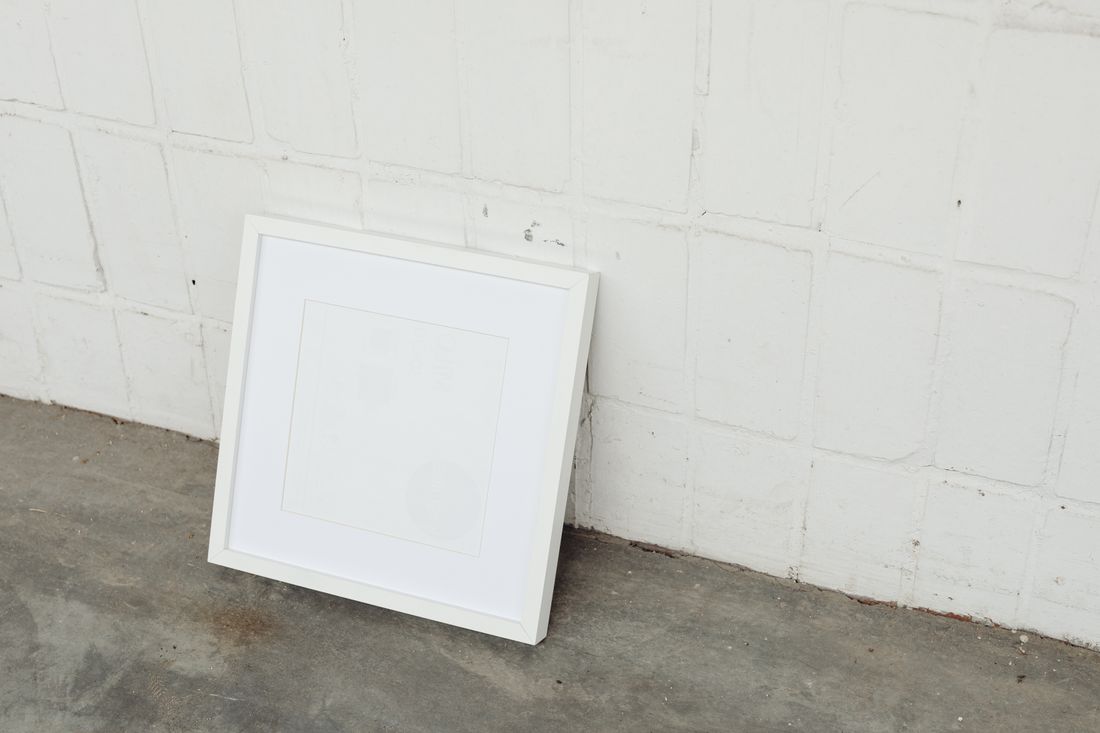 An empty white picture frame with mat leaning against a plain painted brick wall on a concrete floor