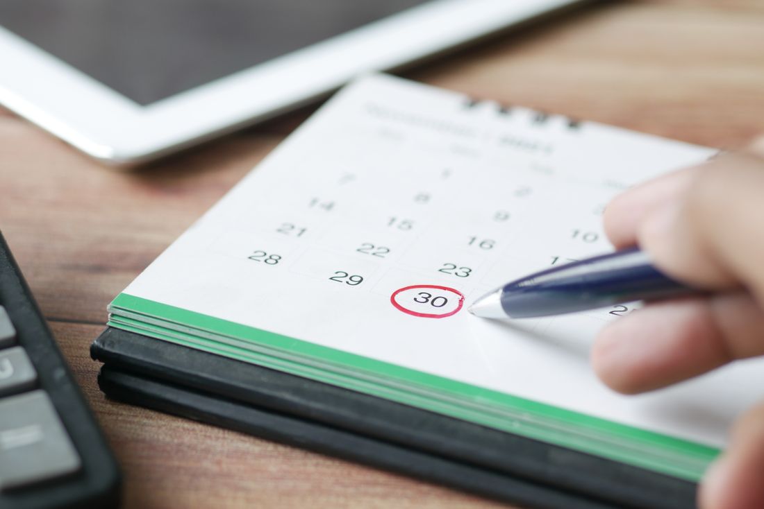 A hand circling a date on a desk calendar with a pen, a tablet and a calculator blurred in the background