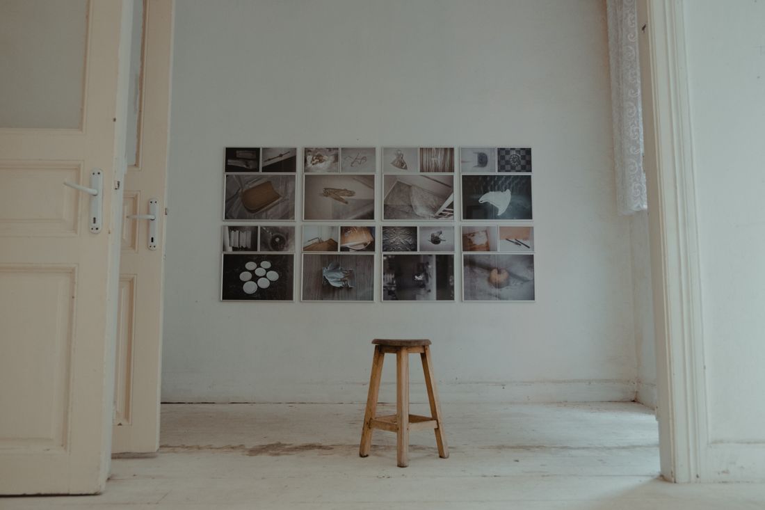 A grid of framed photographs arranged on a gallery wall above a wooden stool in a minimalist room