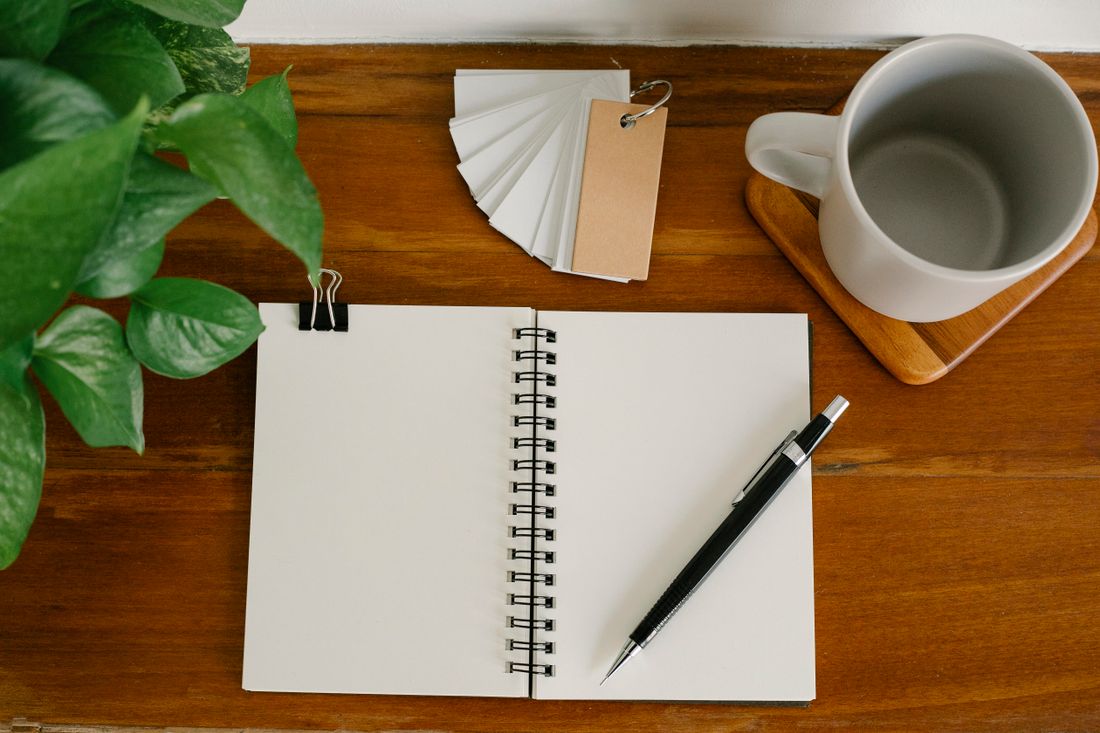 An open blank spiral notebook and a pen on a wooden desk beside a plain mug and a plant